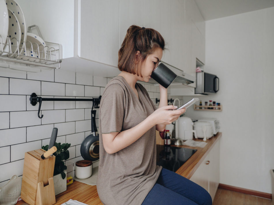 Woman sitting on kitchen counter drinking coffee and scrolling on phone.