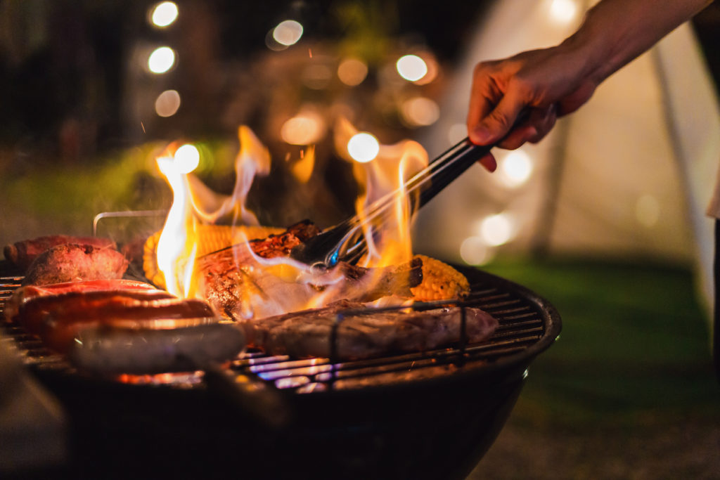 flames leaping up around meat on grill at night, with outdoor lights out of focus in background