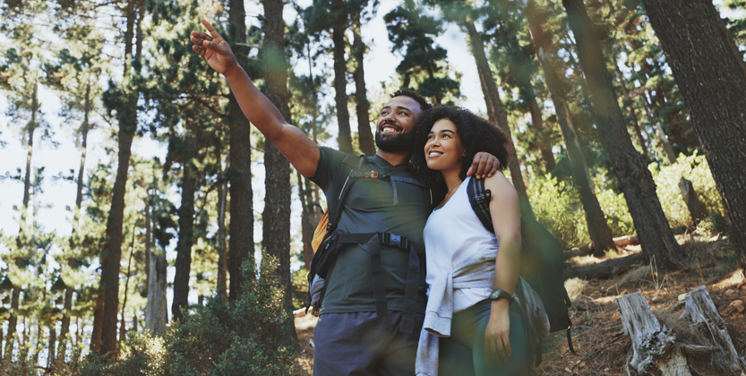 Happy young couple hiking a trail in the forest. Cheerful adventurous man and woman pointing at scenery while taking a break to admire the view and explore their surroundings during a trek in nature